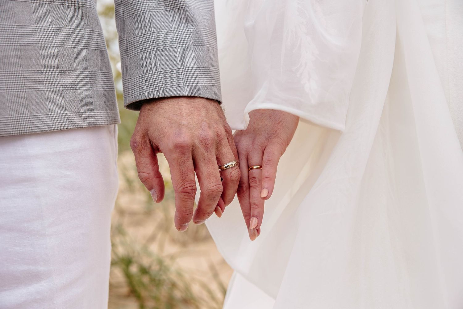 A couple holds hands tenderly while wearing wedding attire, showing rings against a soft blurred background.