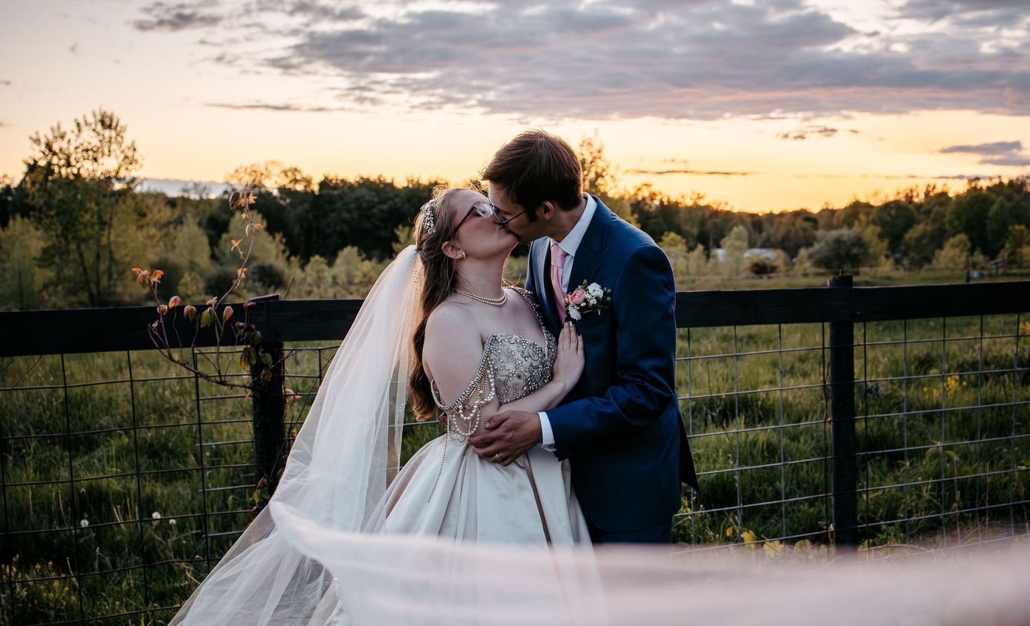A romantic sunset wedding moment captured at a rustic farm with a flowing white veil and navy suit against a pastoral fence.