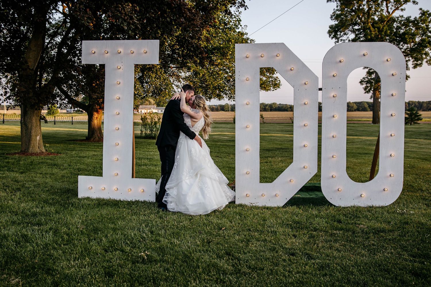 A couple embraces next to large illuminated I DO marquee letters on a grassy lawn at sunset.