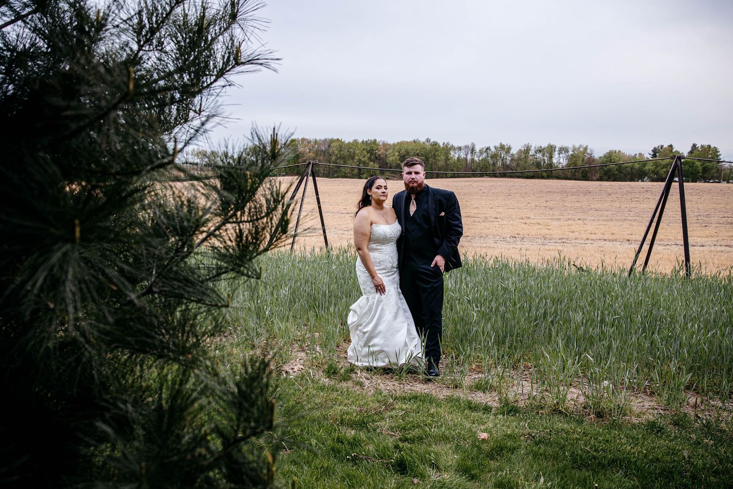 A wedding couple stands together in a grassy field next to farmland on an overcast afternoon.