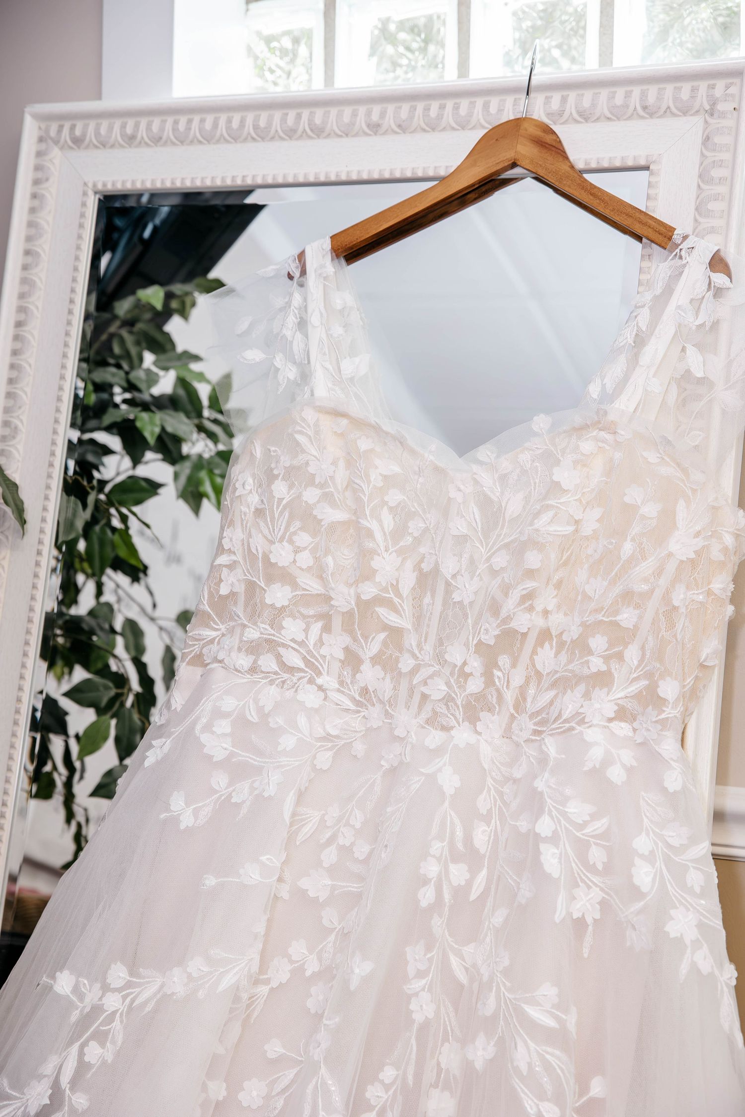 A delicate white wedding dress with floral lace details hangs on a wooden hanger in front of an ornate mirror.