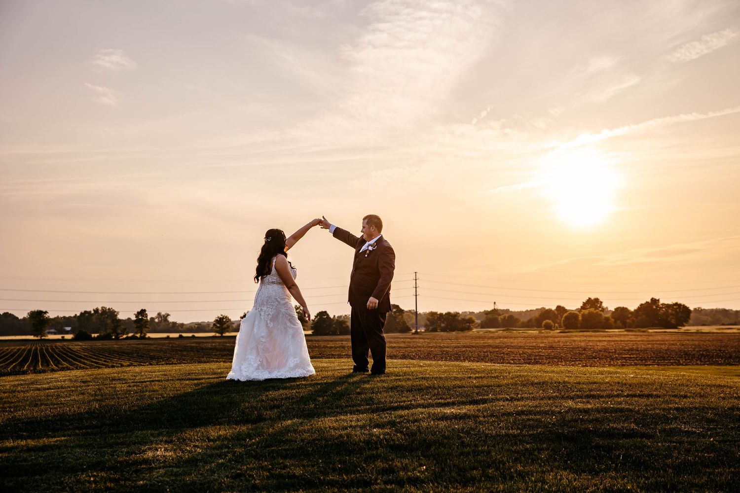 Couple dancing together on a grassy hill during a golden sunset with rural farmland views in the background.