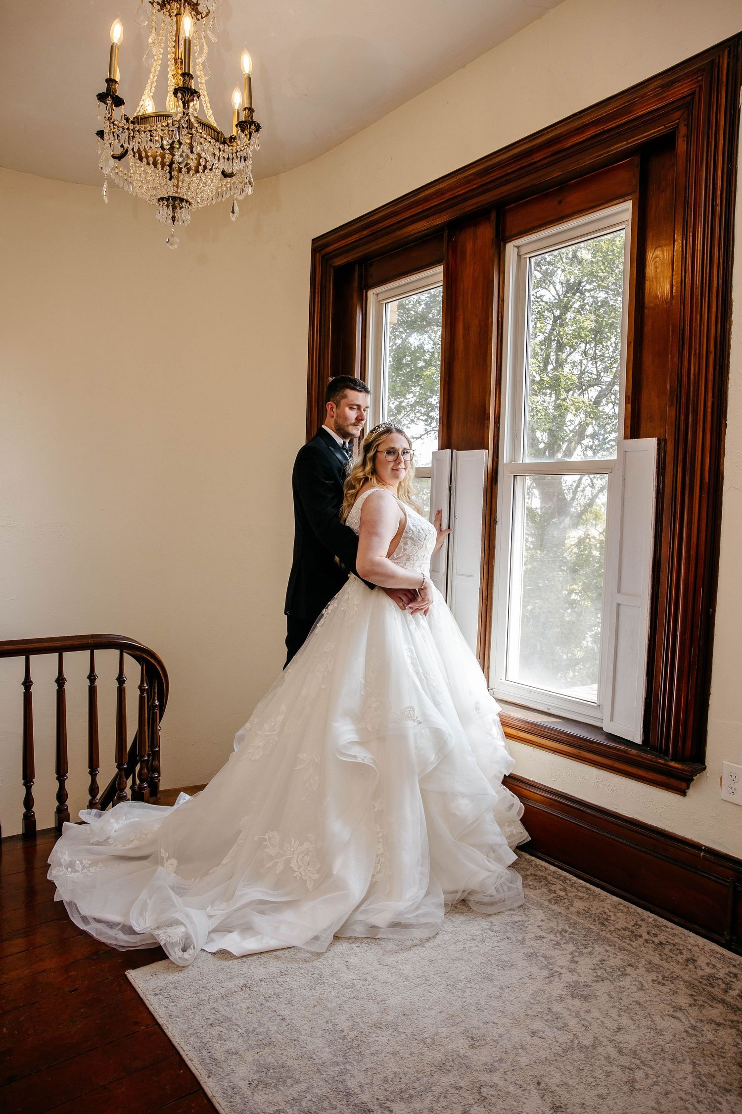 Elegant wedding couple poses by sunlit window in historic venue with ornate chandelier and rich wood trim.