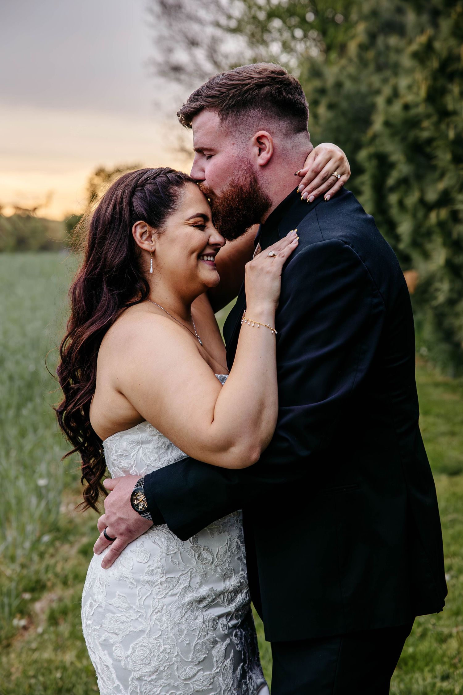 A romantic wedding couple embraces in a garden at sunset, with the bride in a lace gown and groom in a black suit.