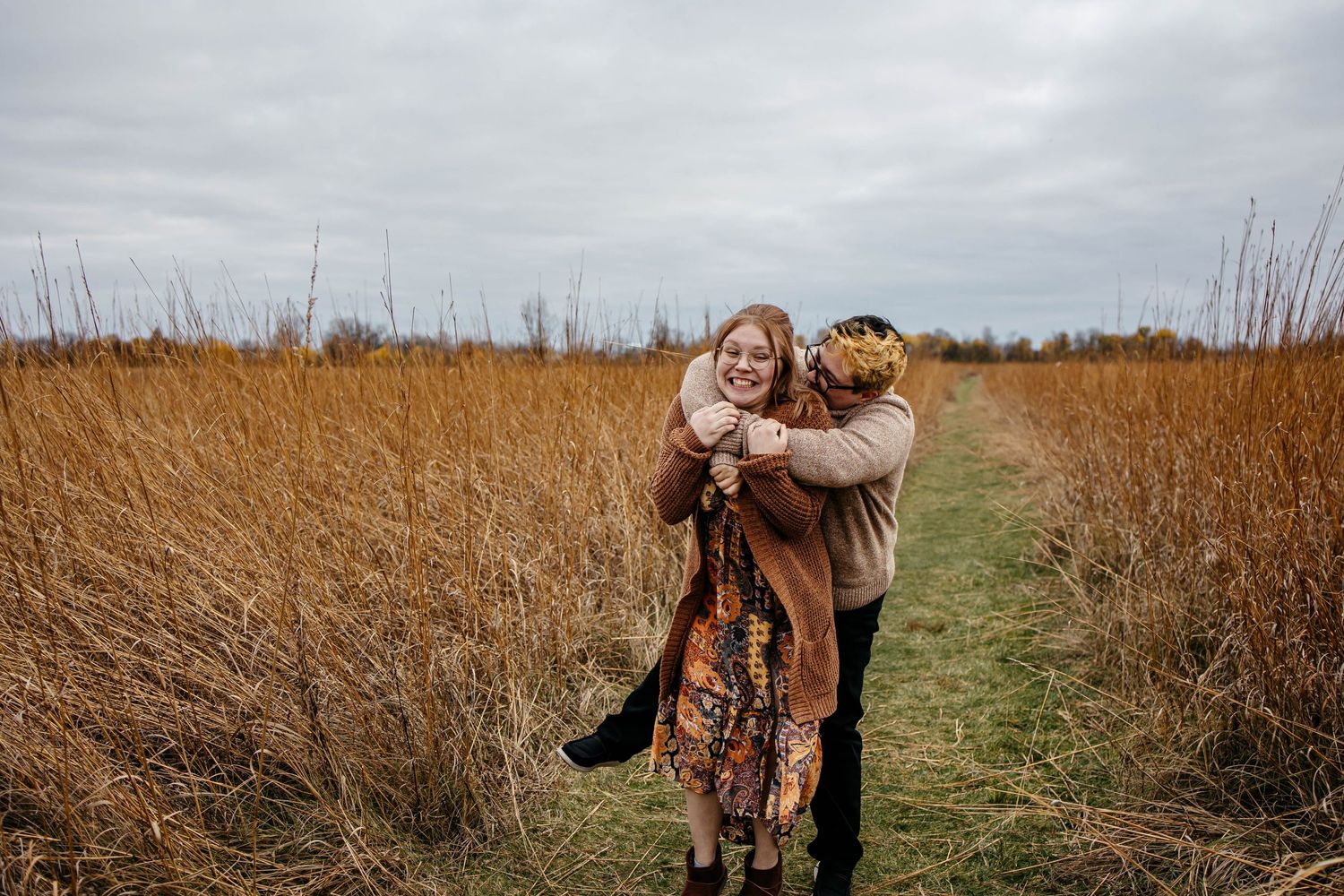 A candid moment between two people embracing on a pathway through tall golden grass on an overcast autumn day.