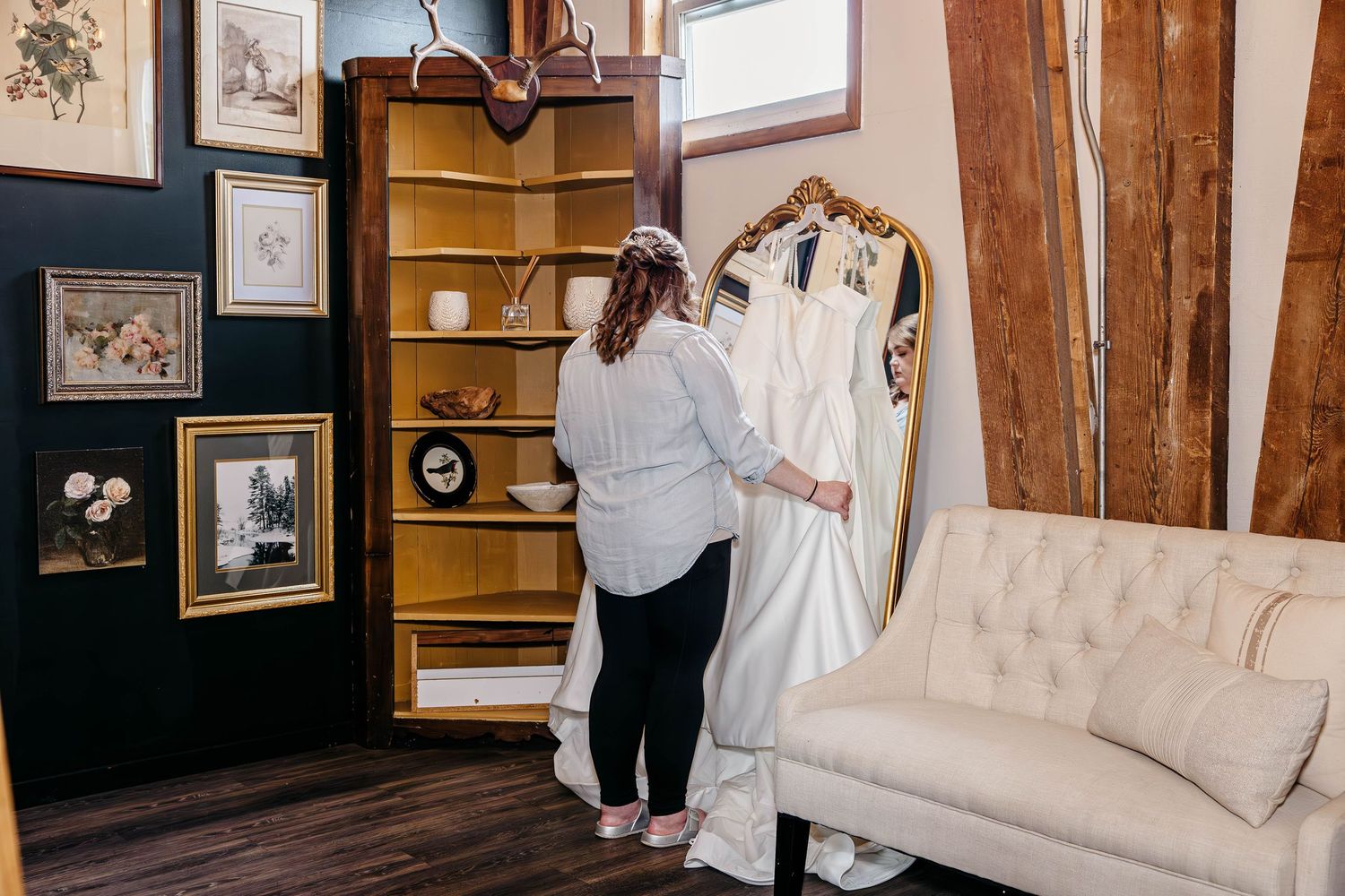 Someone looks at a white wedding dress hanging on display next to a wooden bookshelf and beige couch in a rustic room.