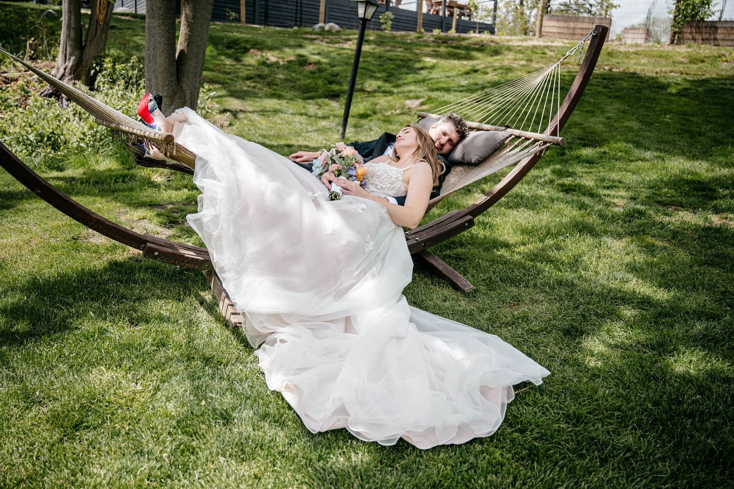 A couple relaxes together in a hammock outdoors, with a flowing white wedding dress draped over the side.