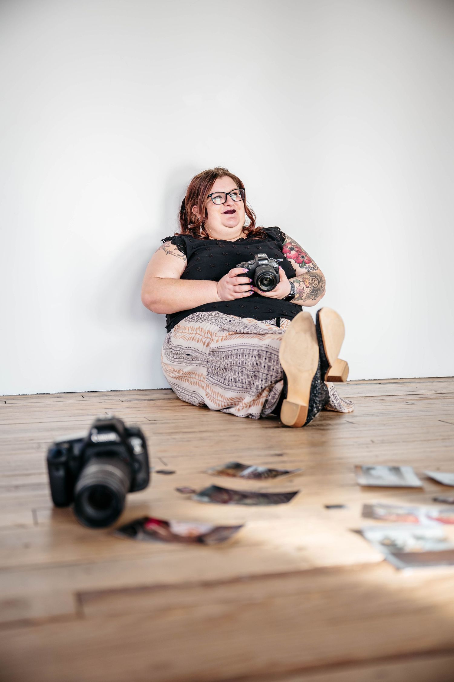 Photographer sitting on wooden floor with DSLR camera and scattered photos around while laughing joyfully.