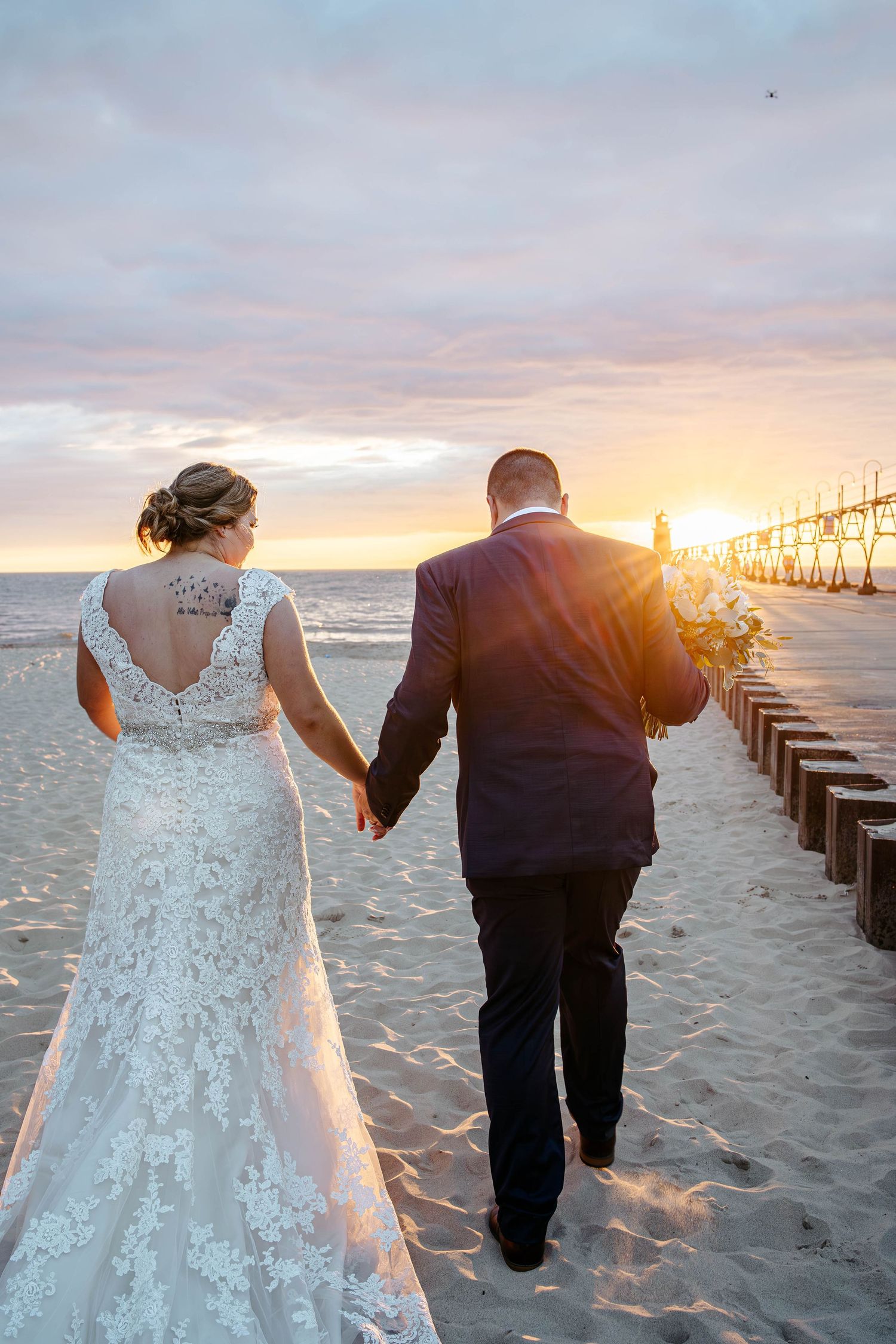 Newlywed couple walking on beach at sunset near wooden pier as golden light illuminates their romantic moment.