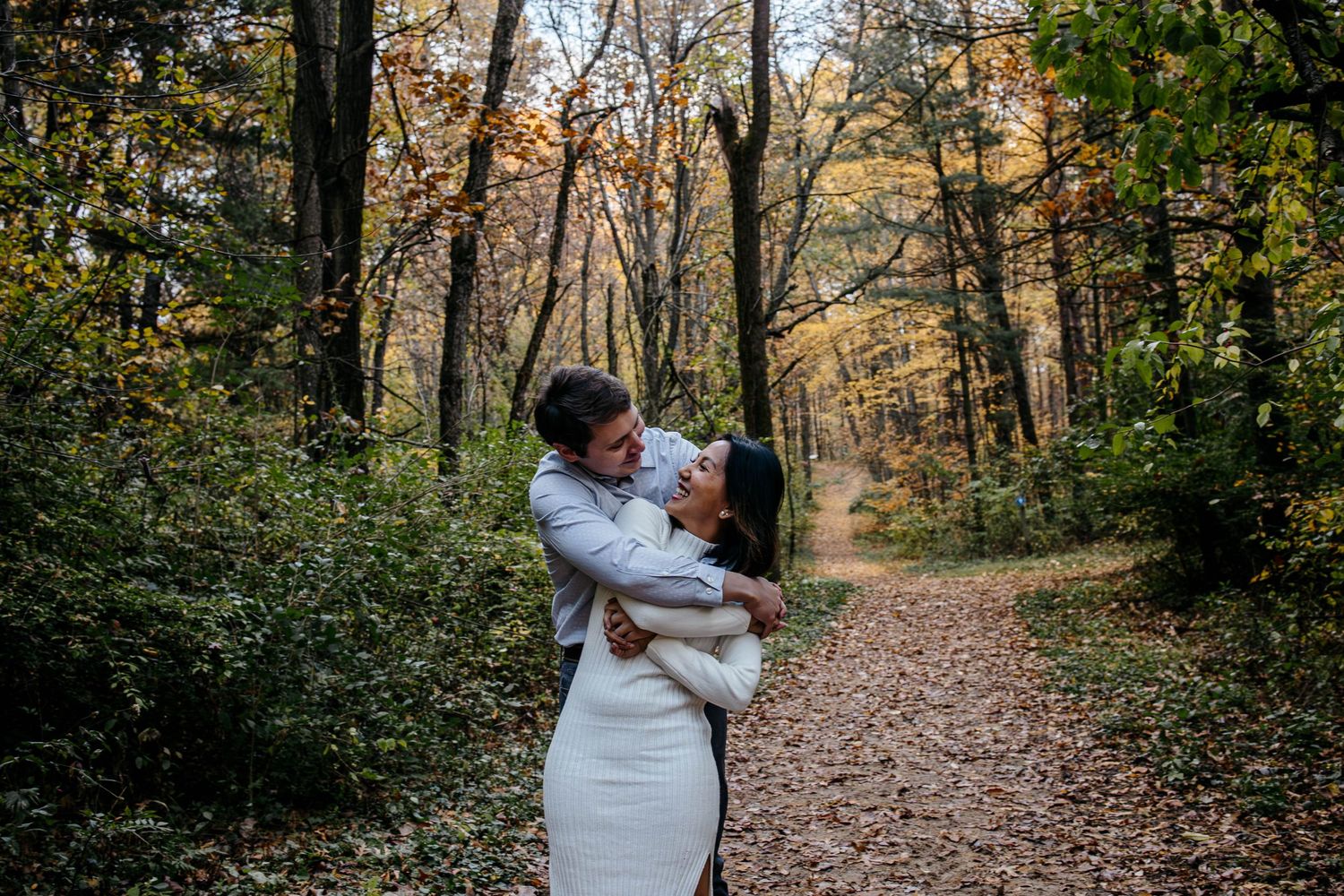 A couple embraces on a tree-lined autumn path covered in fallen leaves during golden hour.