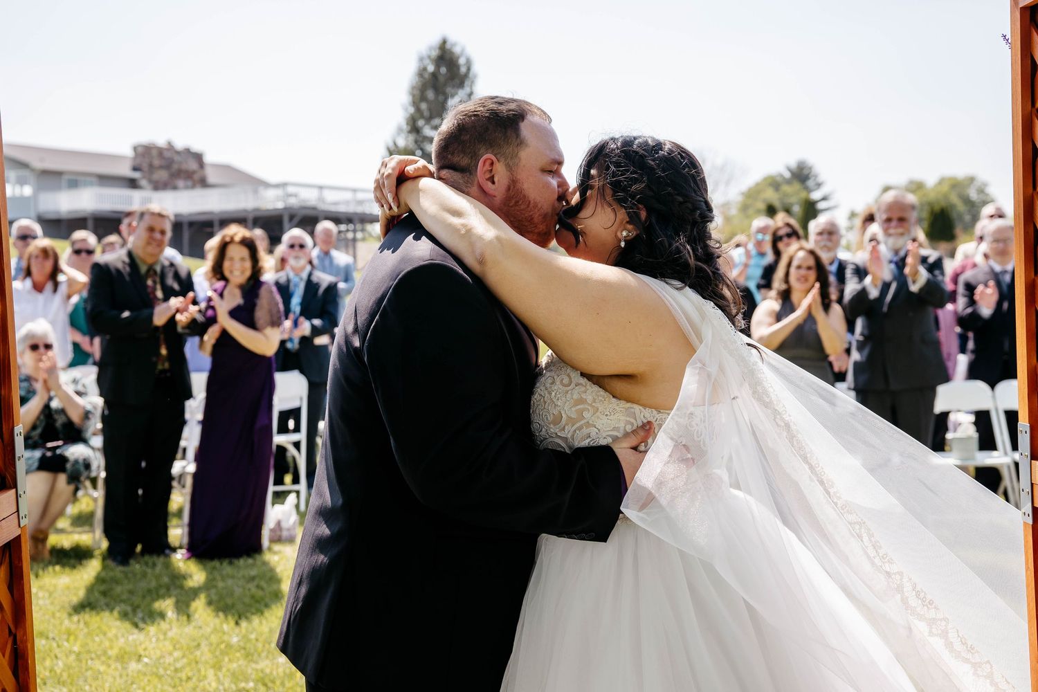 A newlywed couple embraces in a romantic kiss at their outdoor wedding ceremony as guests clap and celebrate.