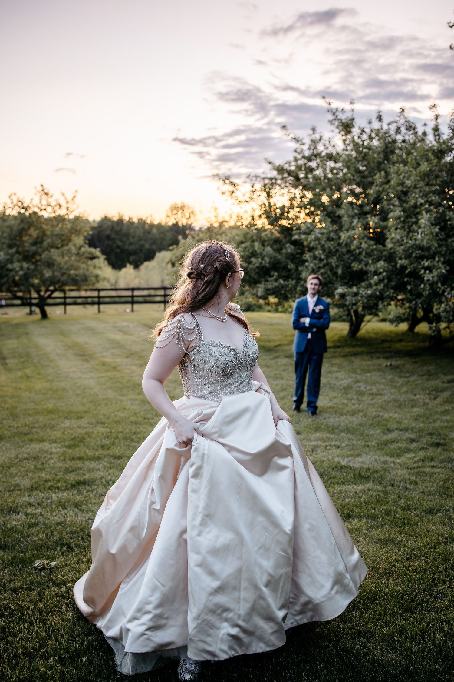A romantic sunset wedding photo featuring a bride in a flowing white ballgown dress standing in an orchard at dusk.