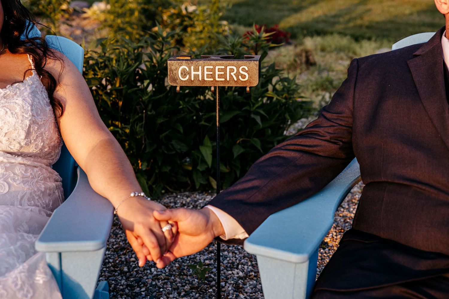Two newlyweds hold hands while sitting beneath a rustic wooden 'Cheers' sign at an outdoor wedding reception.