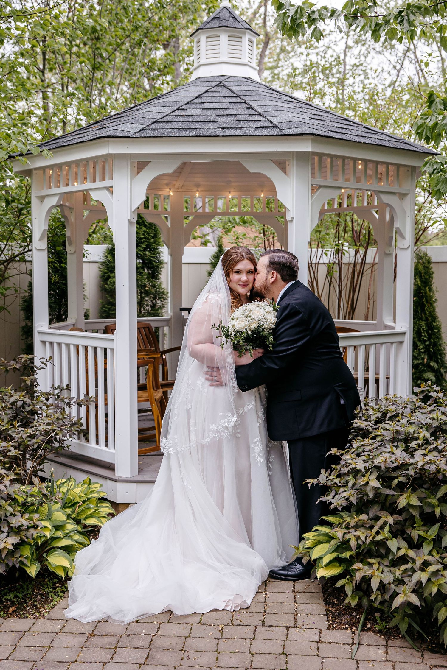 A romantic couple share a tender moment in a white garden gazebo surrounded by lush greenery and stone paving.