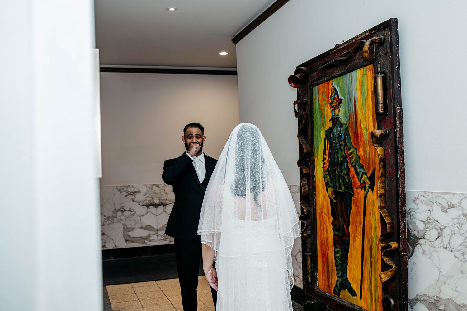 A formal wedding moment captured in an elegant room with marble walls and a vibrant orange artwork displayed.