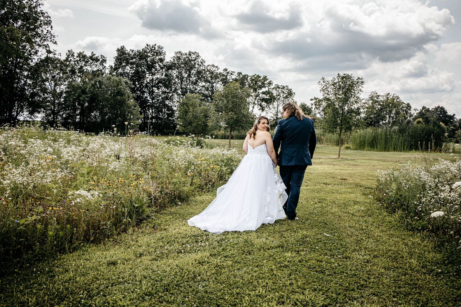 A couple in wedding attire stands in a scenic meadow with wildflowers and trees on a cloudy summer day.