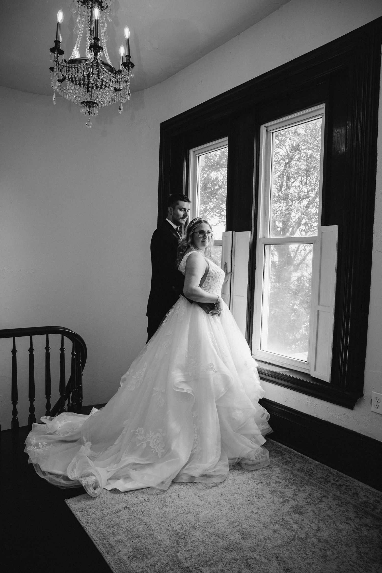 A black and white photo of newlyweds posing by tall windows under an elegant chandelier.
