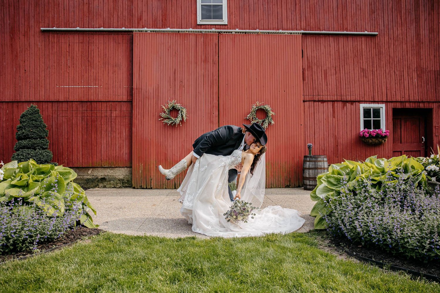 A couple shares a romantic dip dance move in front of a rustic red barn with decorative garden landscaping.
