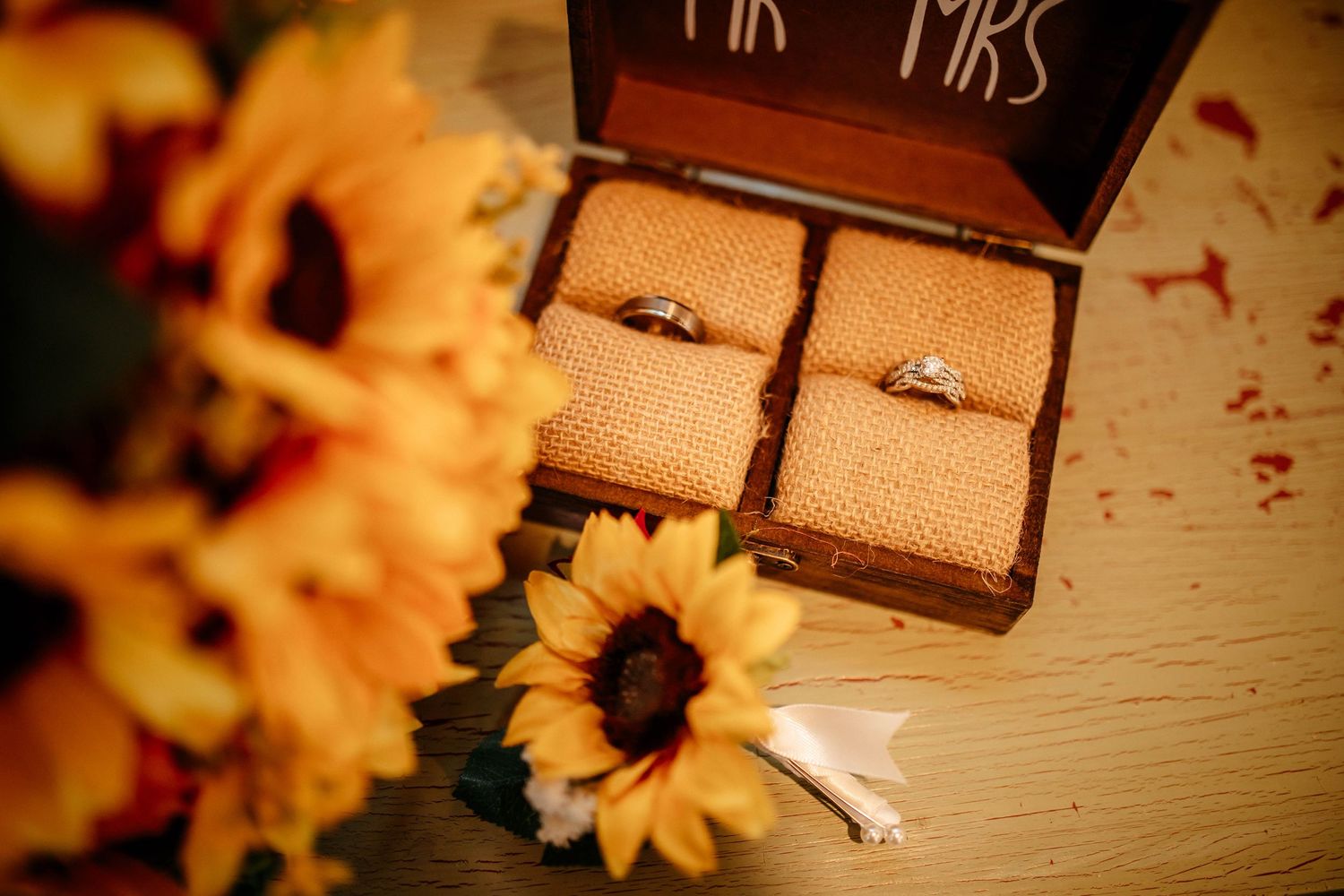 Two wedding rings displayed in a wooden box next to yellow sunflowers on a white surface with romantic lighting.