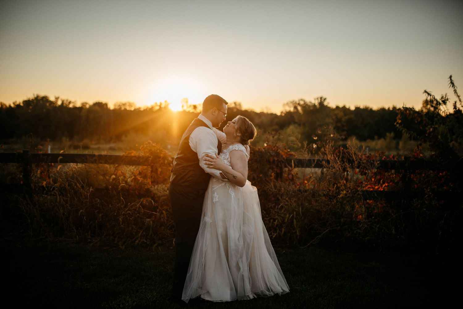 A couple share a romantic embrace at sunset in a rustic outdoor setting with golden light streaming through the trees.