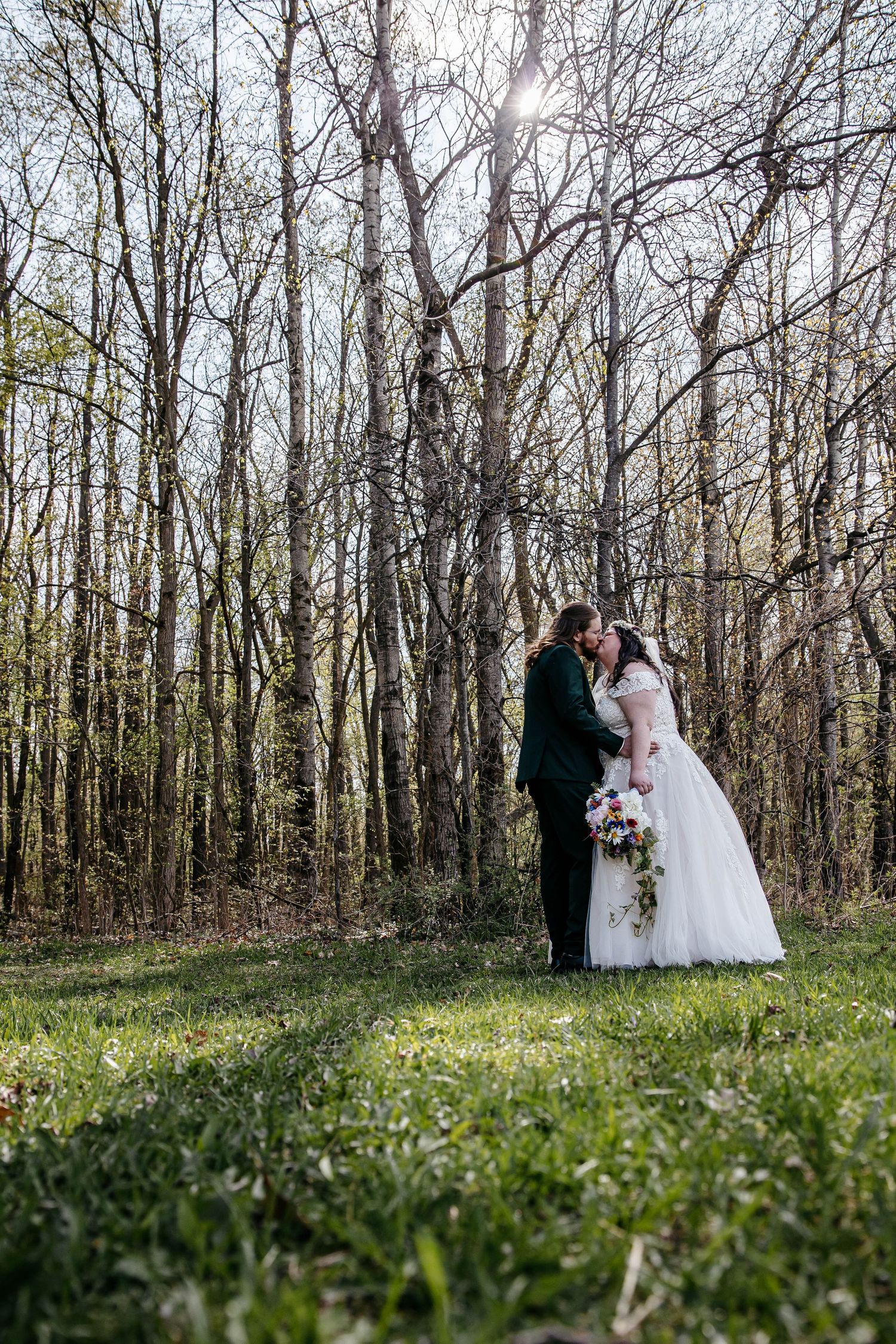 A wedding couple shares a tender moment in a scenic woodland setting with bare trees and sunlight filtering through branches.