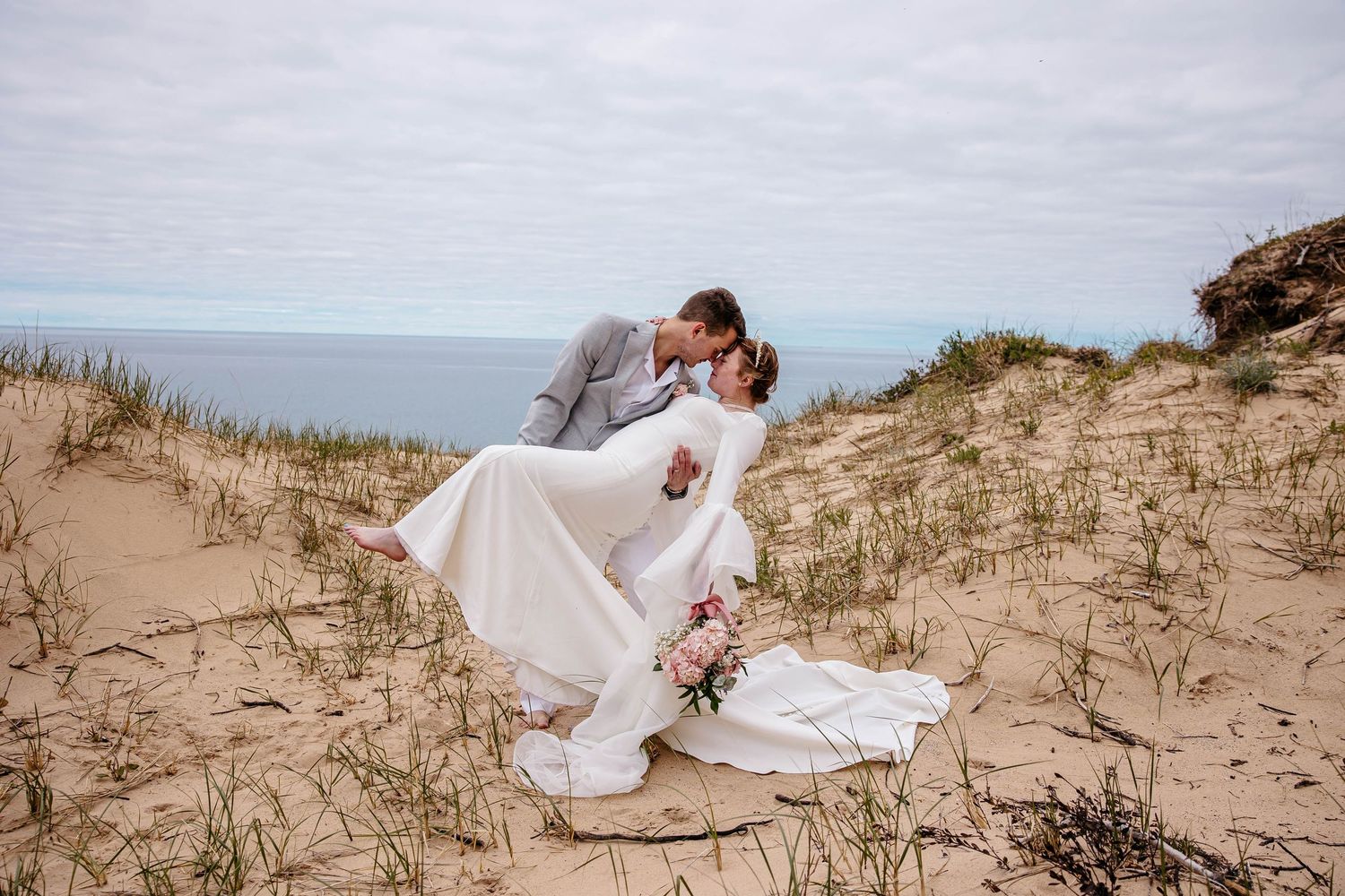 A beach wedding couple reclines across a series of dunes along a romantic sandy shoreline at sunset.
