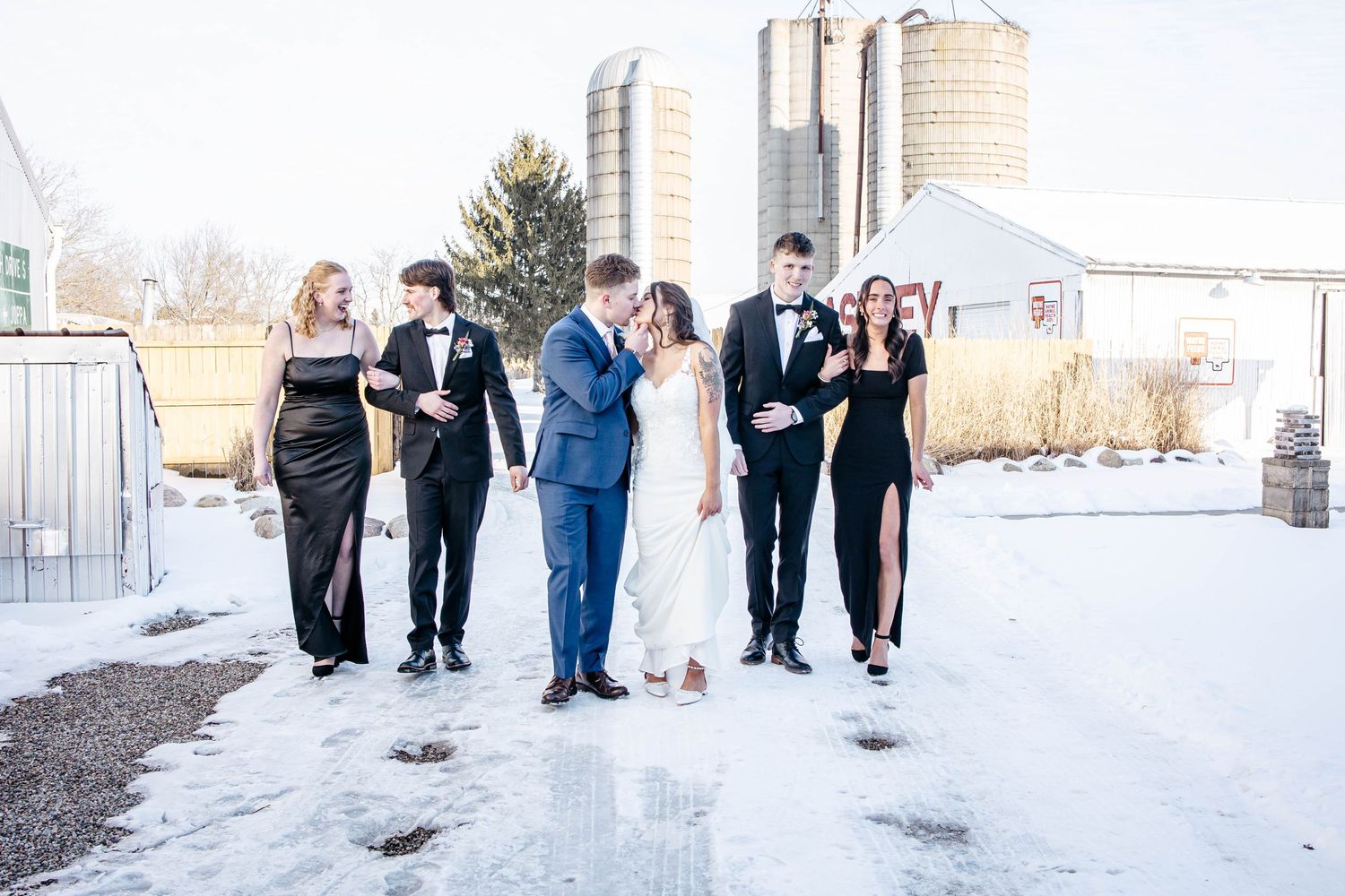 A wedding party in formal attire stands together in the snow with farm silos visible in the background.