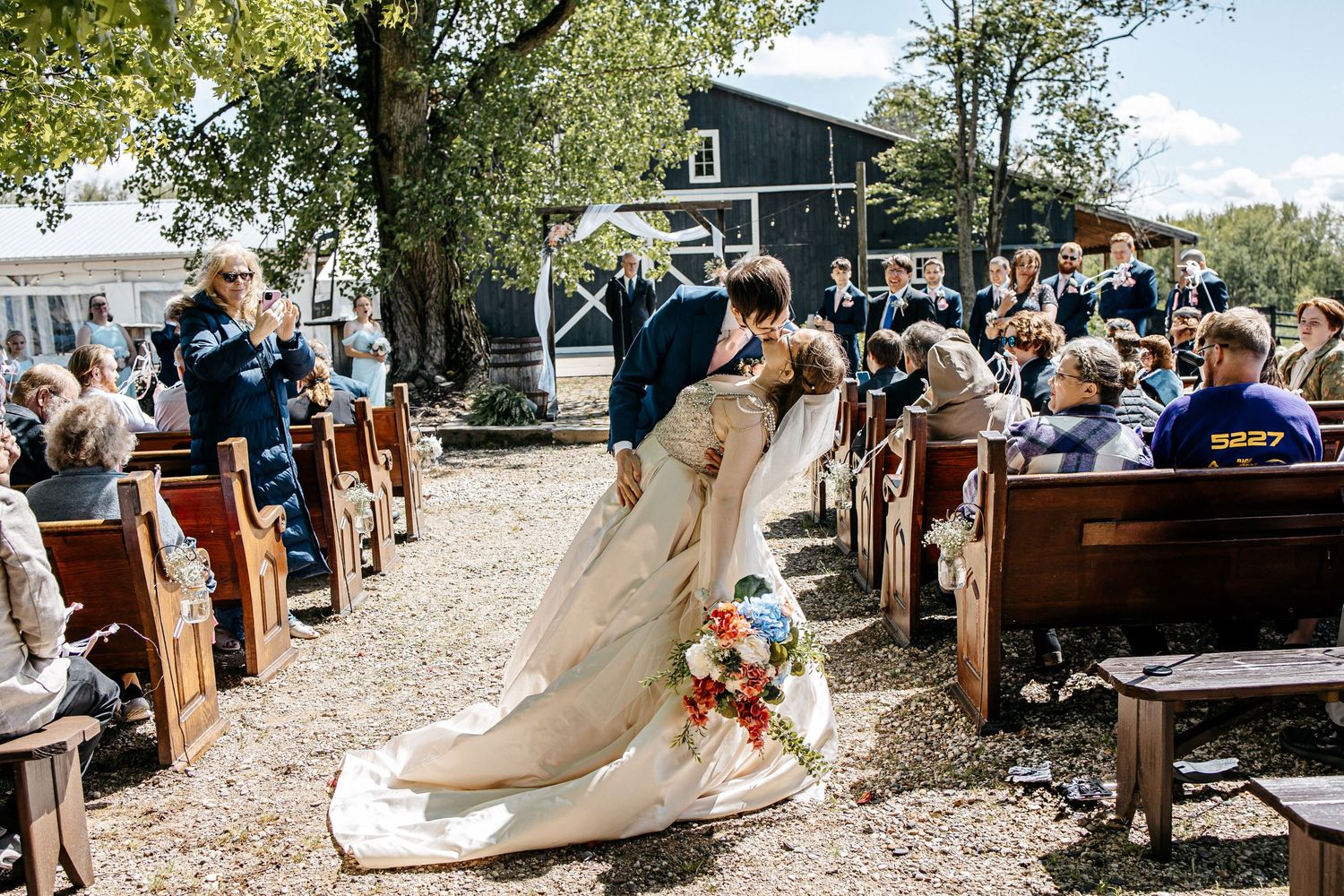 An outdoor wedding ceremony takes place on a gravel path with wooden pew seating under trees next to a blue house.