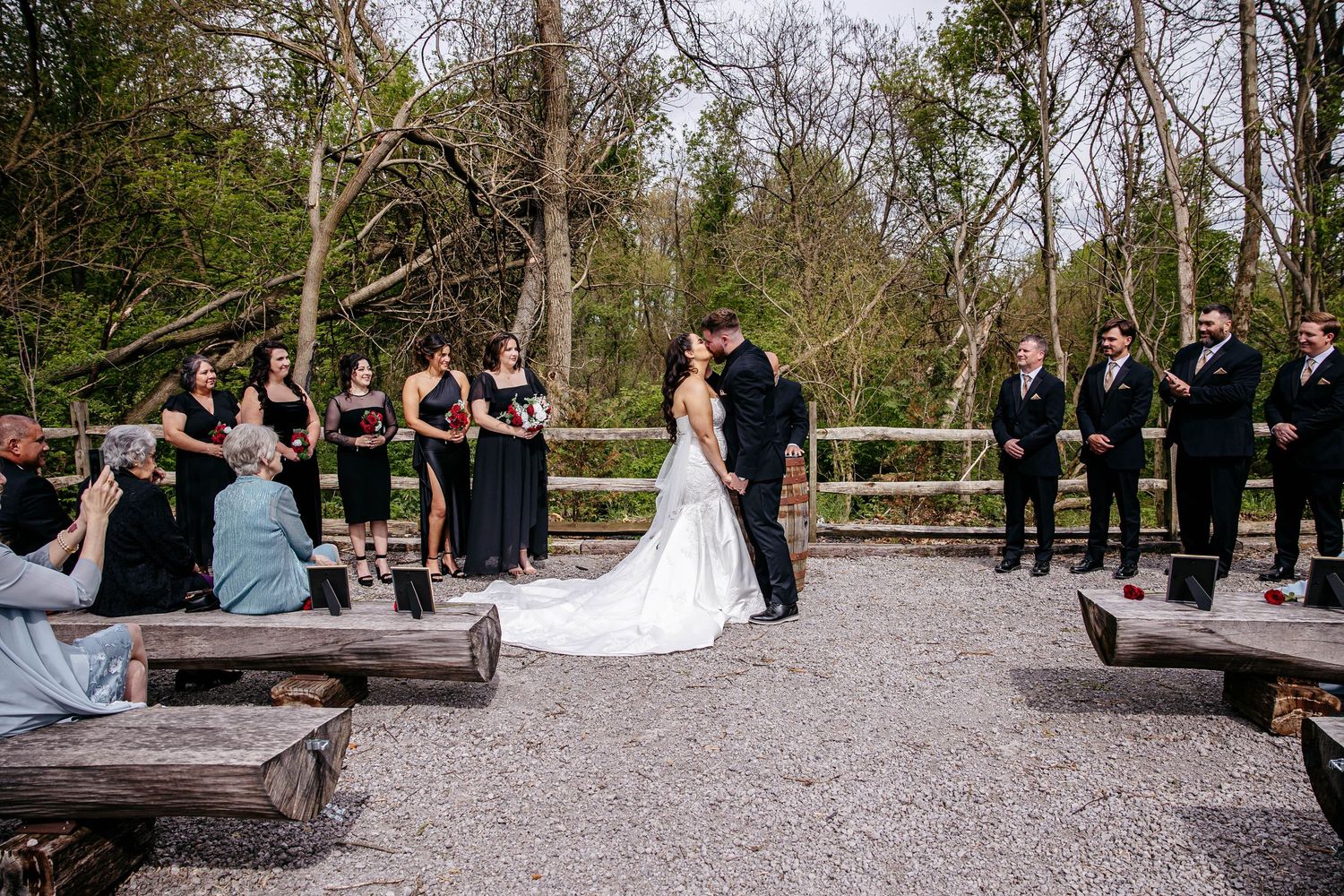 Wedding ceremony at an outdoor venue with rustic wooden benches and lush forest backdrop.