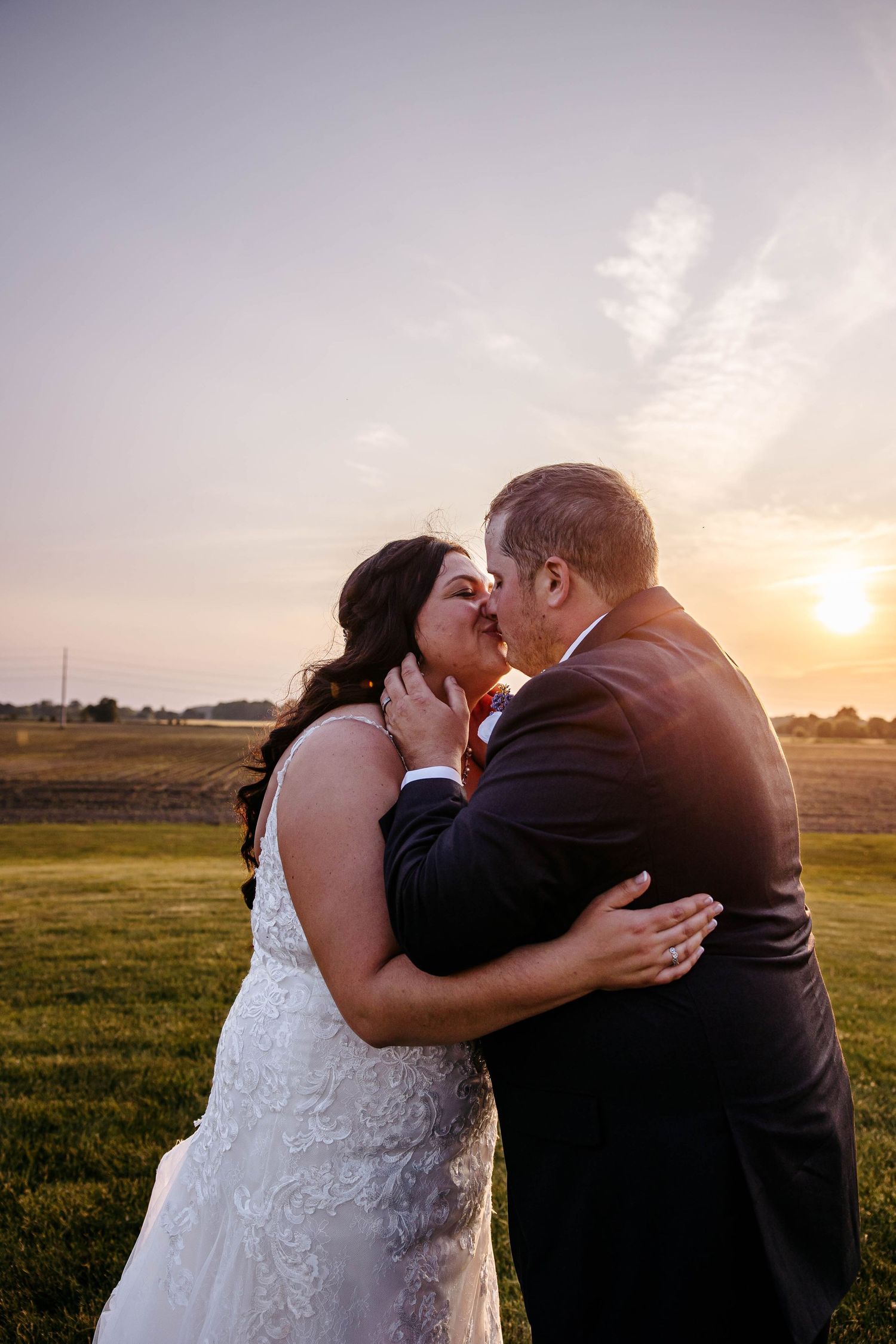 A couple in wedding attire share a romantic kiss at sunset in a grassy field.