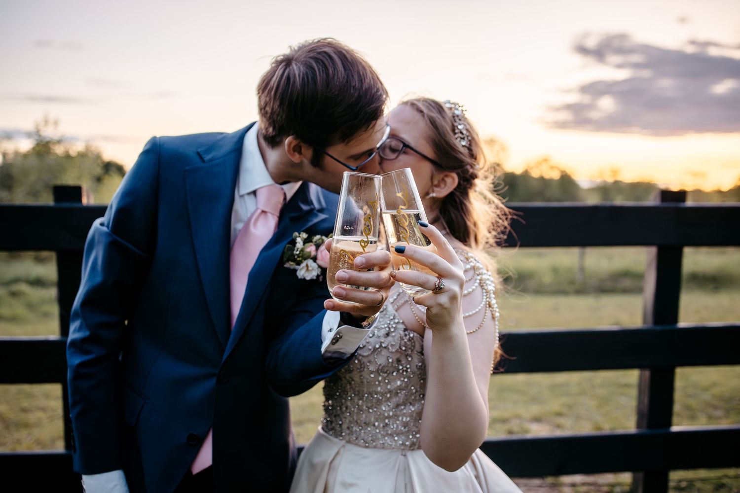 A bride and groom in formal attire share a romantic toast with champagne glasses at sunset by a wooden fence.