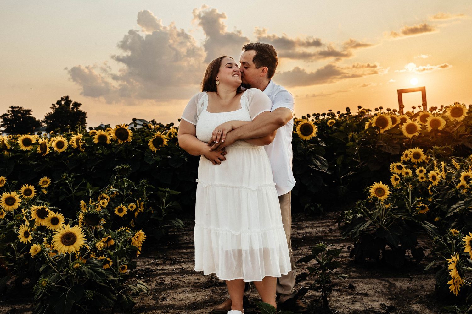 A couple embraces in a sunflower field at golden hour sunset with warm glowing light in the background.