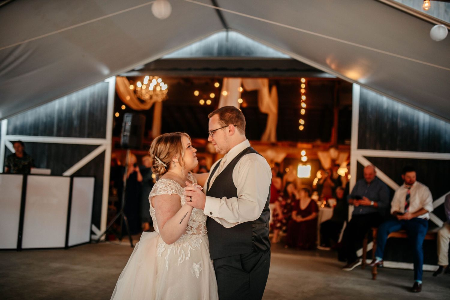 A couple shares their first dance at a wedding reception under a white tent with warm lighting and string lights.