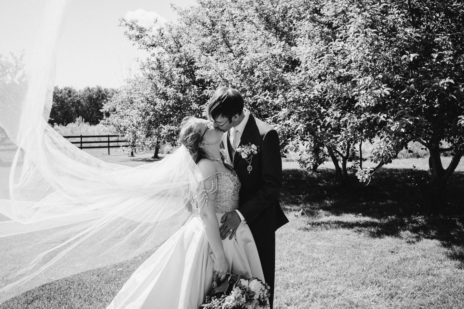 Black and white romantic wedding photo showing couple embracing outdoors as brides veil flows in the wind.