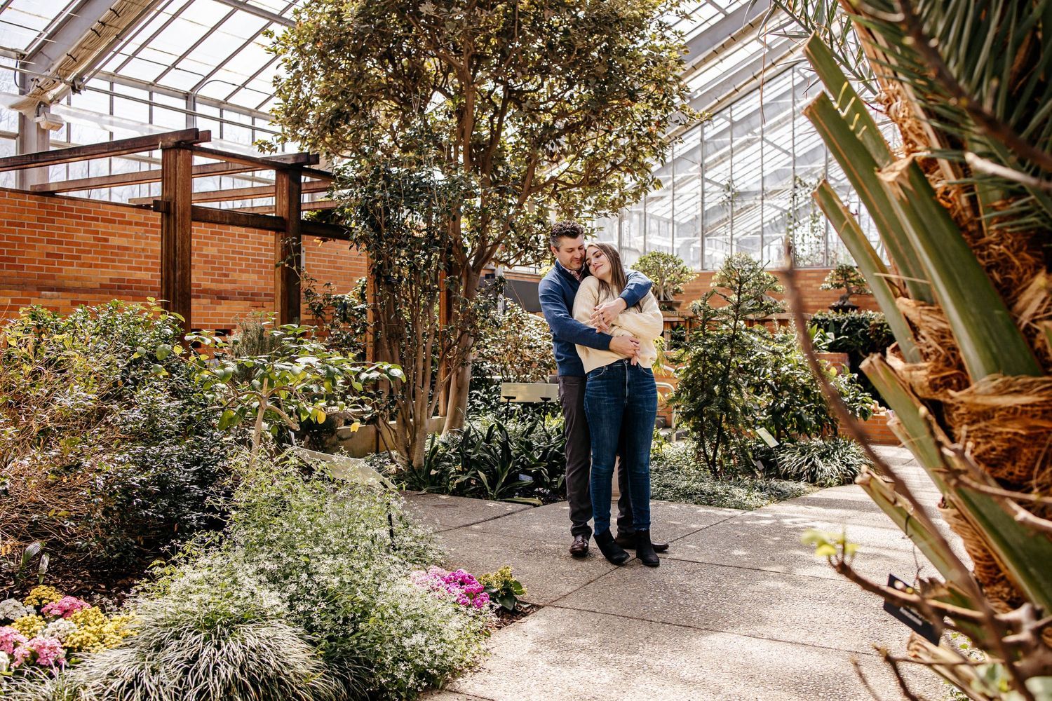 A couple embraces in a sunlit greenhouse filled with lush plants, flowers and a glass ceiling overhead.
