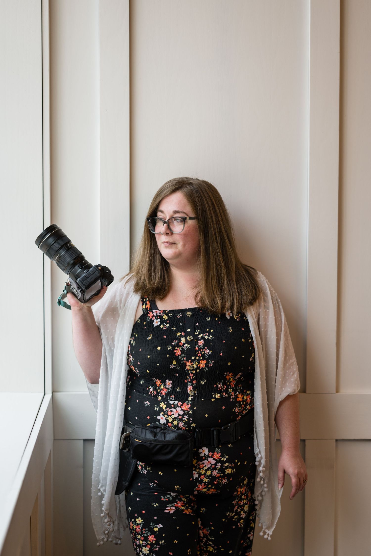 A photographer in a floral black dress takes different playful poses with their camera against a light wall.