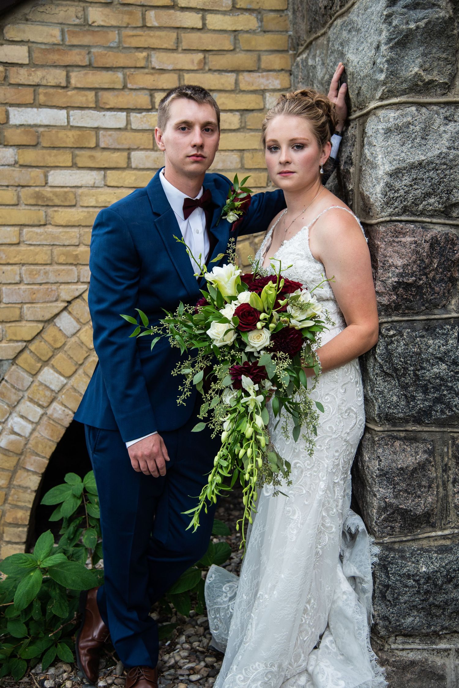 Wedding couple poses against brick wall with elegant white and burgundy floral bouquet.