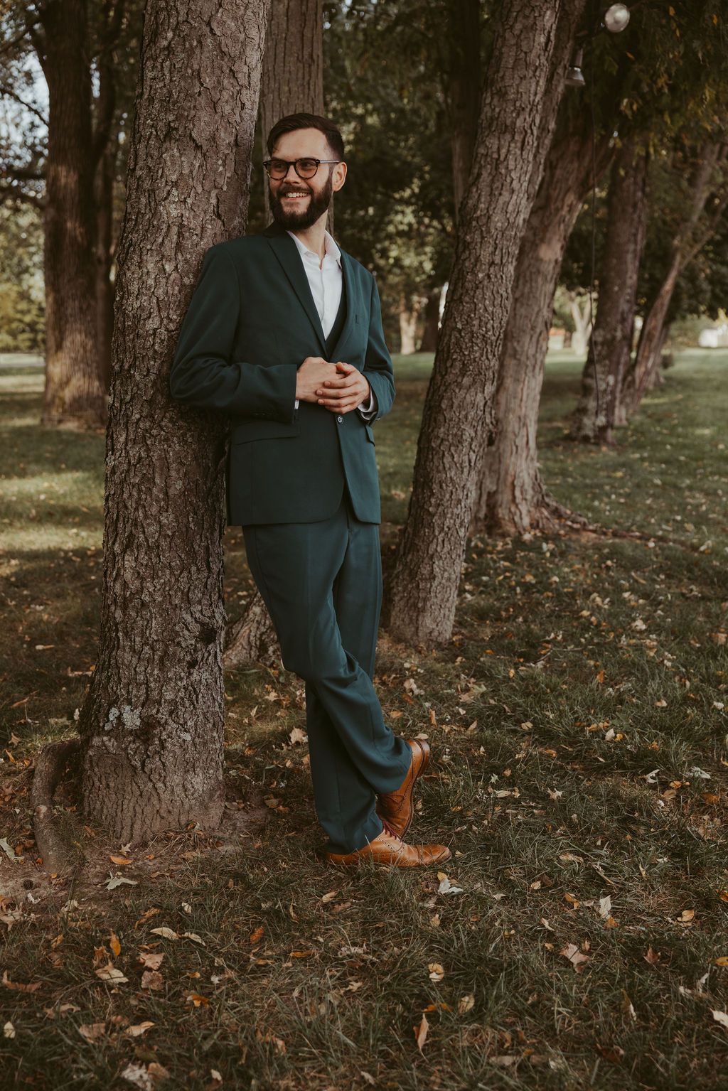 A person in a dark green suit and glasses leans casually against a tree trunk in an autumn forest setting.