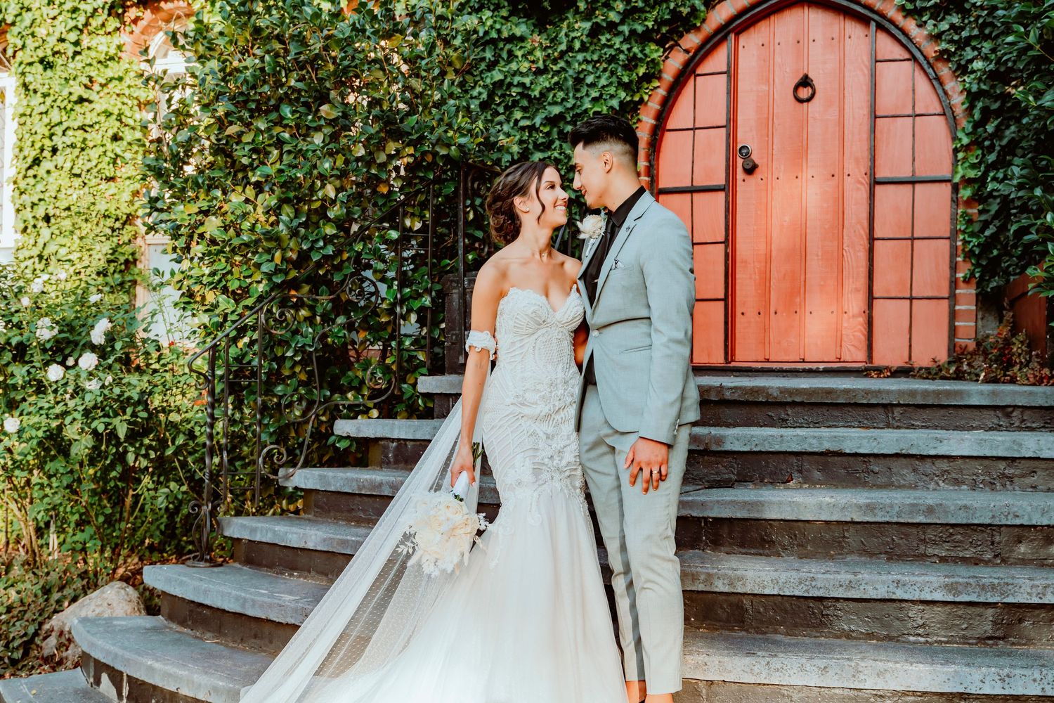 A couple in elegant wedding attire poses together on stone steps in front of an arched red wooden door surrounded by greenery.