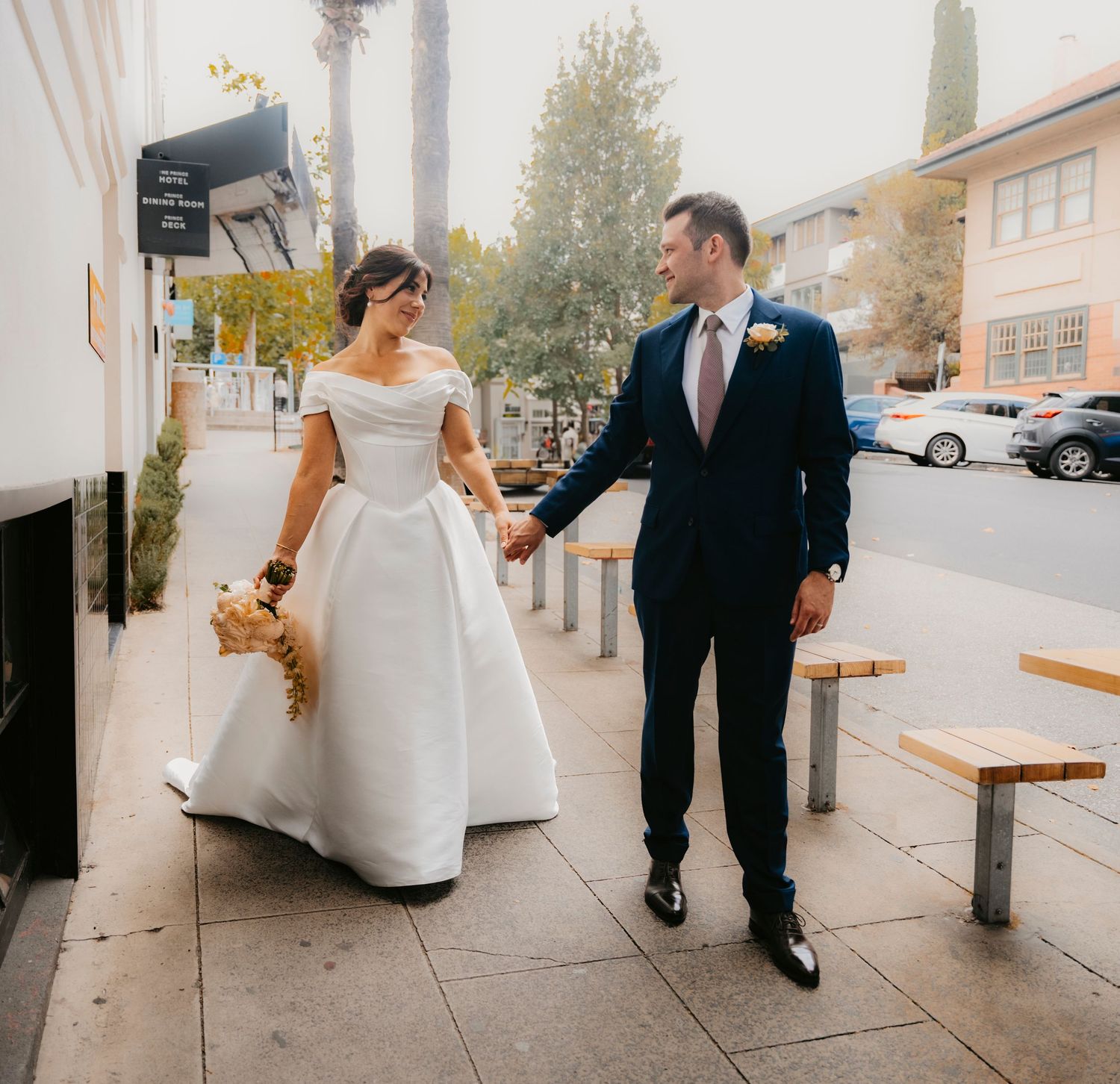 A bride and groom walking together on a city sidewalk on their wedding day, she in white and he in a dark suit.