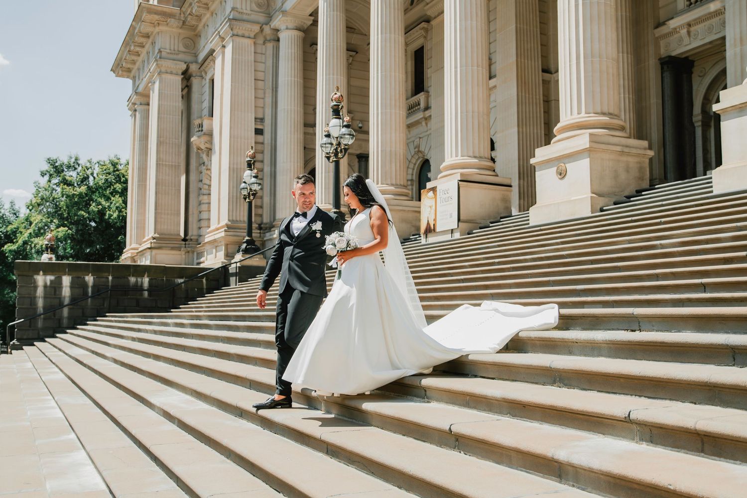 A couple in wedding attire descending grand stone steps with neoclassical columns at an elegant historic building.