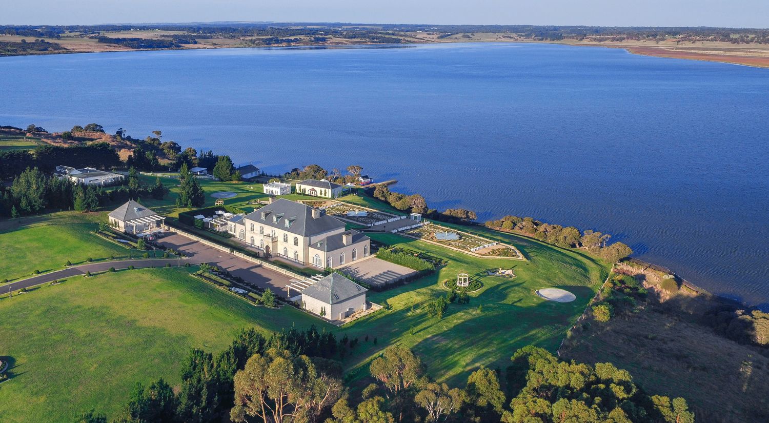 Aerial view of a grand historic mansion and manicured grounds situated along a scenic blue coastal waterfront.