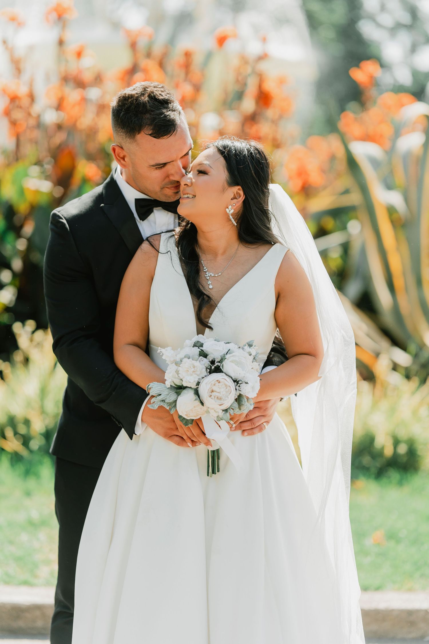 A romantic wedding moment captured outdoors with a bridal bouquet of white flowers and orange floral garden backdrop.