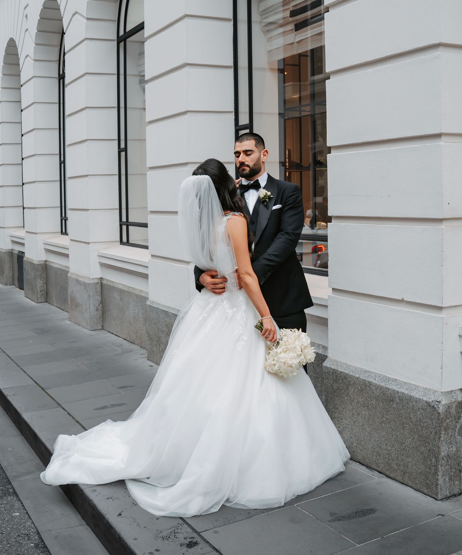 A romantic wedding couple share an intimate kiss on a city street in front of a white stone building.