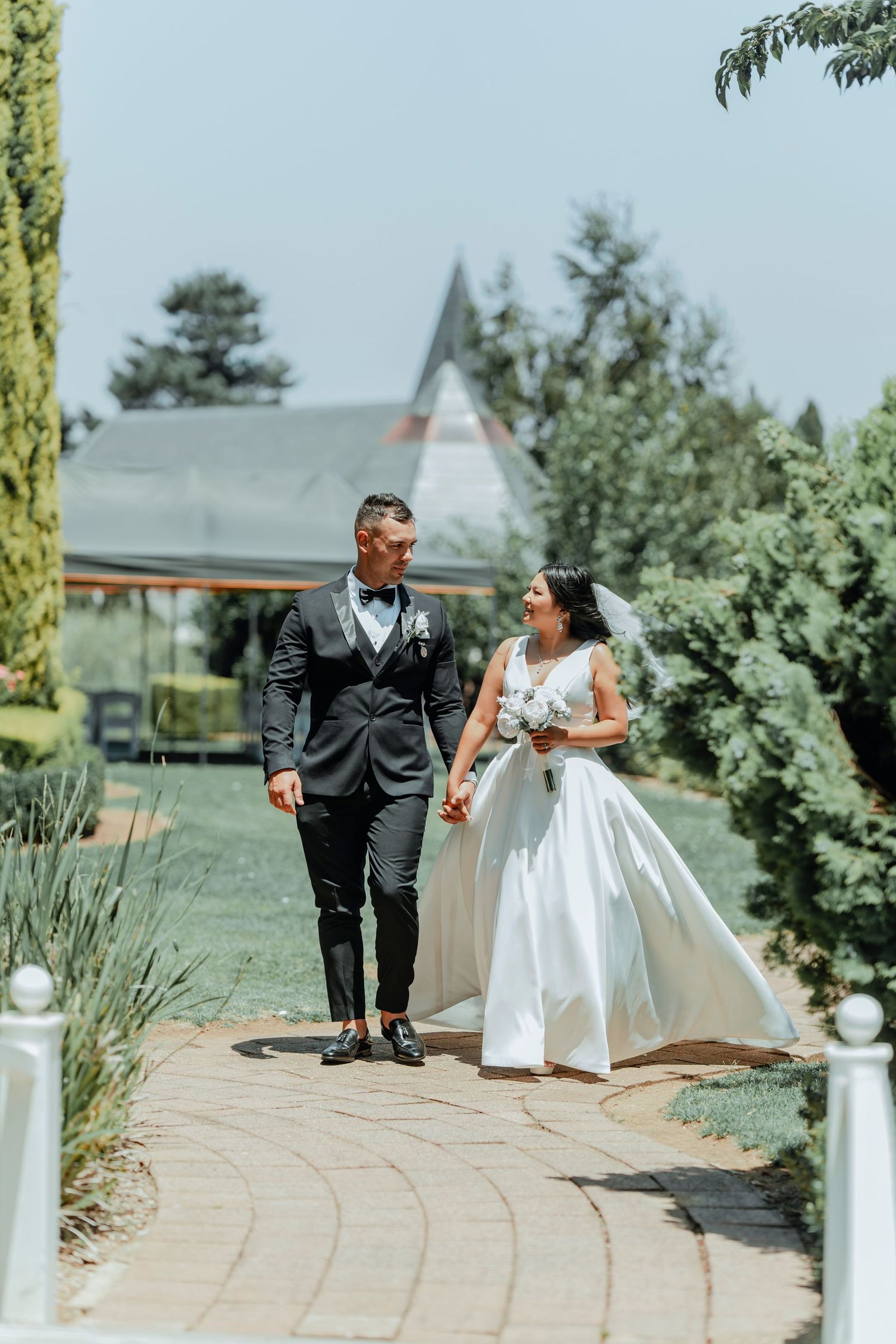 Newlywed couple walking together on curved garden path with chapel in background on sunny summer day.
