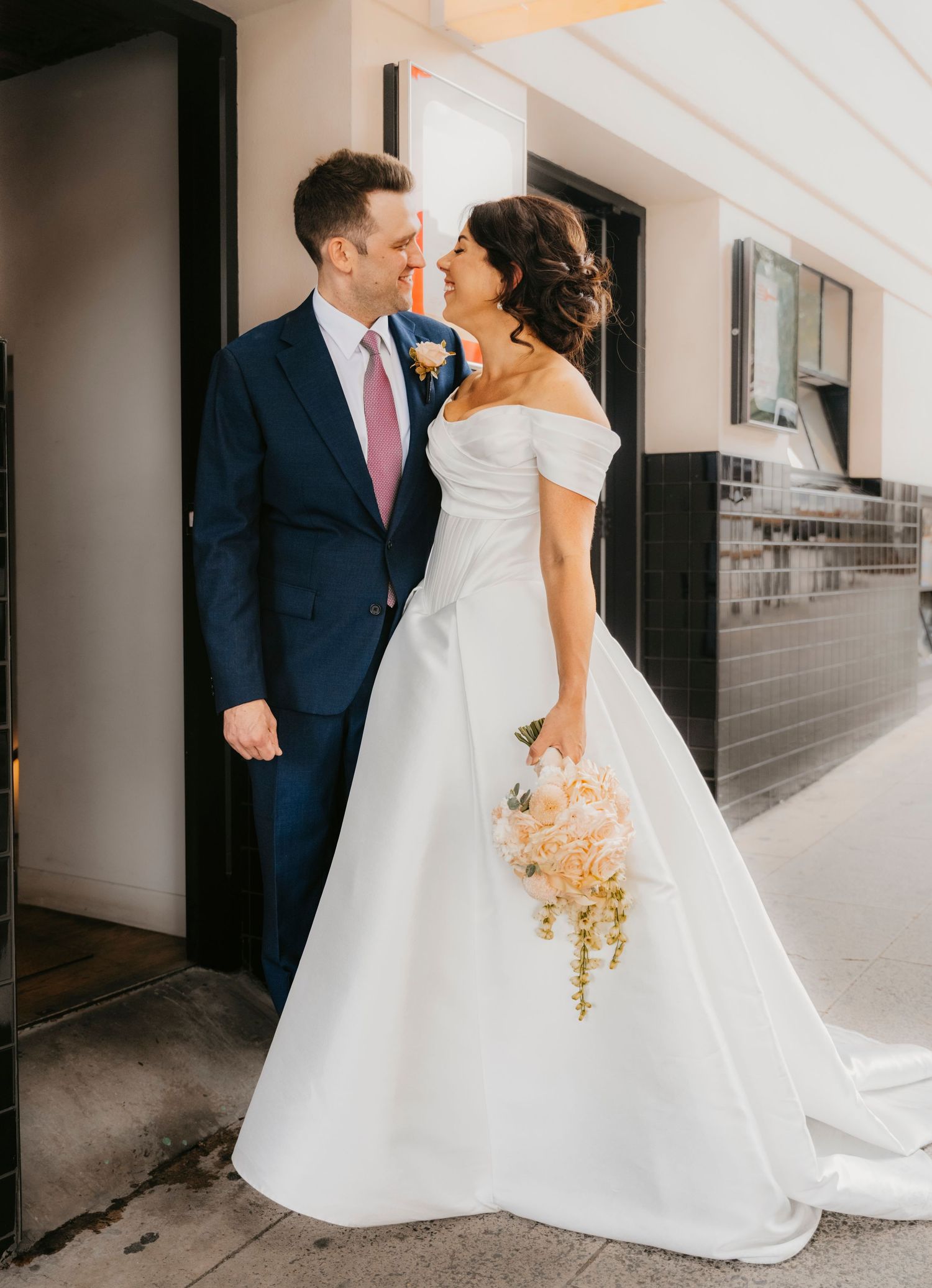 A couple in formal wedding attire share an intimate moment while standing in a modern hallway with blush florals.