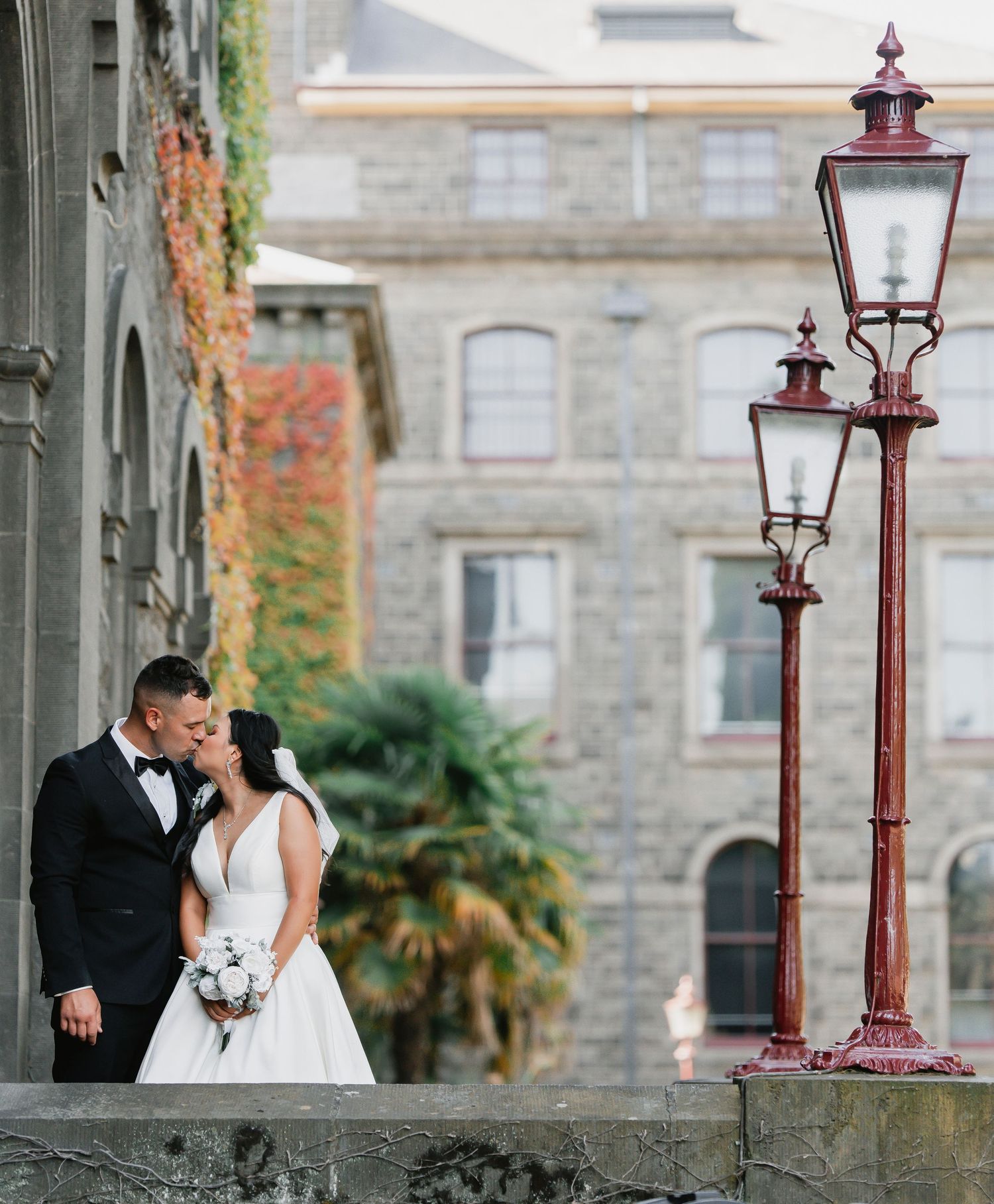 A wedding couple embraces near vintage-style street lamps outside a stone building with autumn foliage.