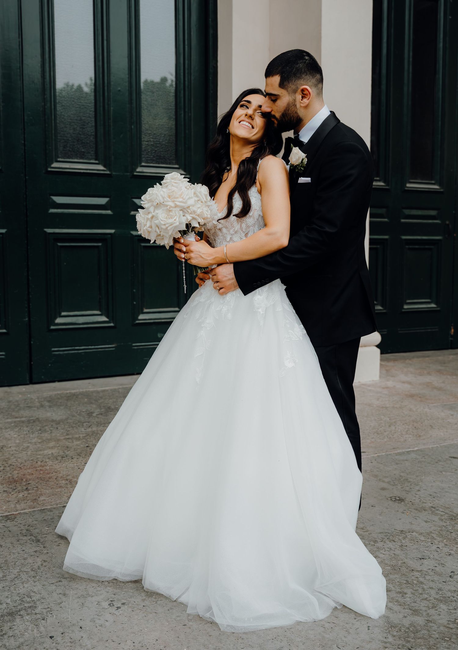 A newlywed couple embraces romantically in front of dark green doors while the bride holds a white floral bouquet.