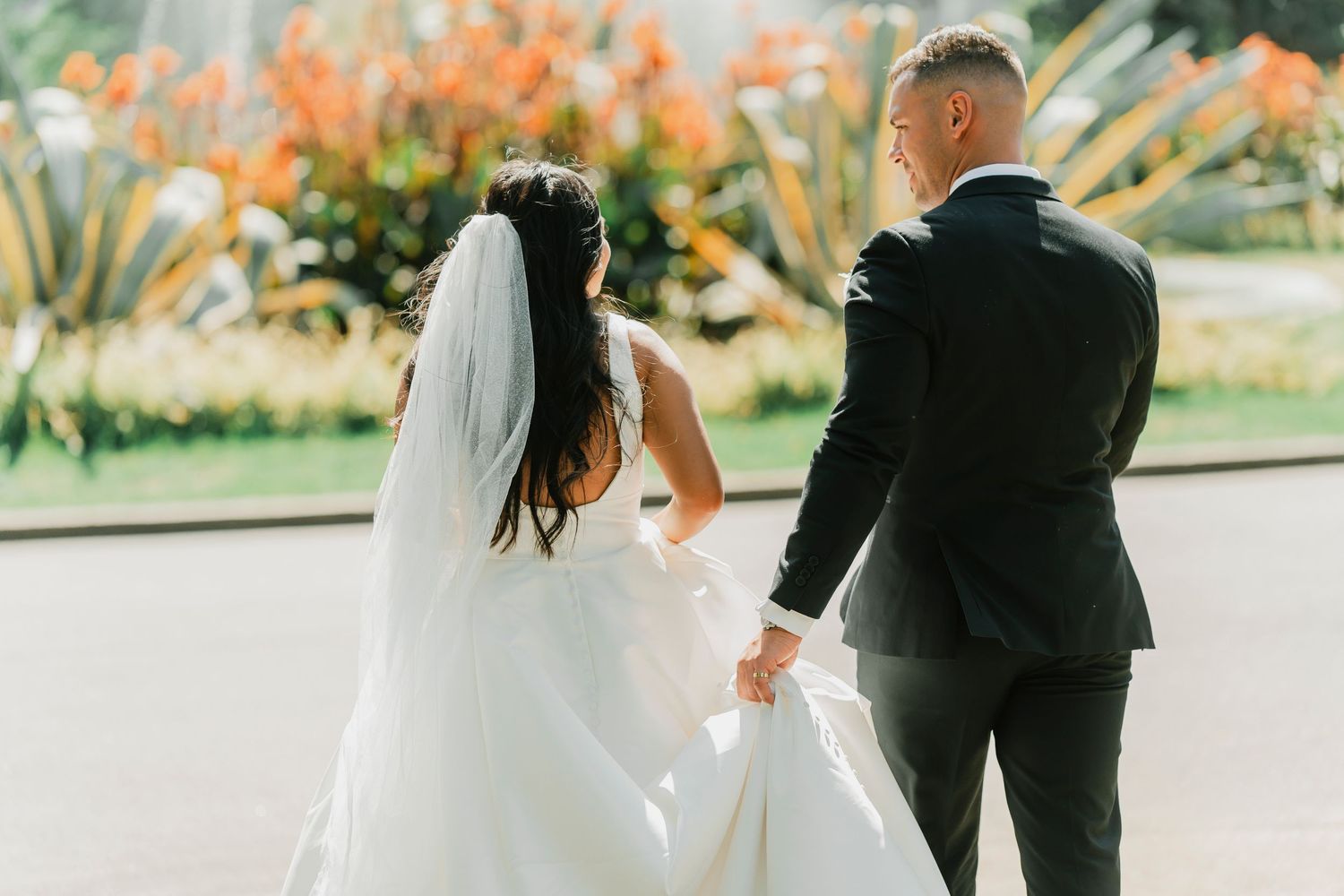 A bride in a white gown and groom in black suit walking together outdoors with orange flowers in background.