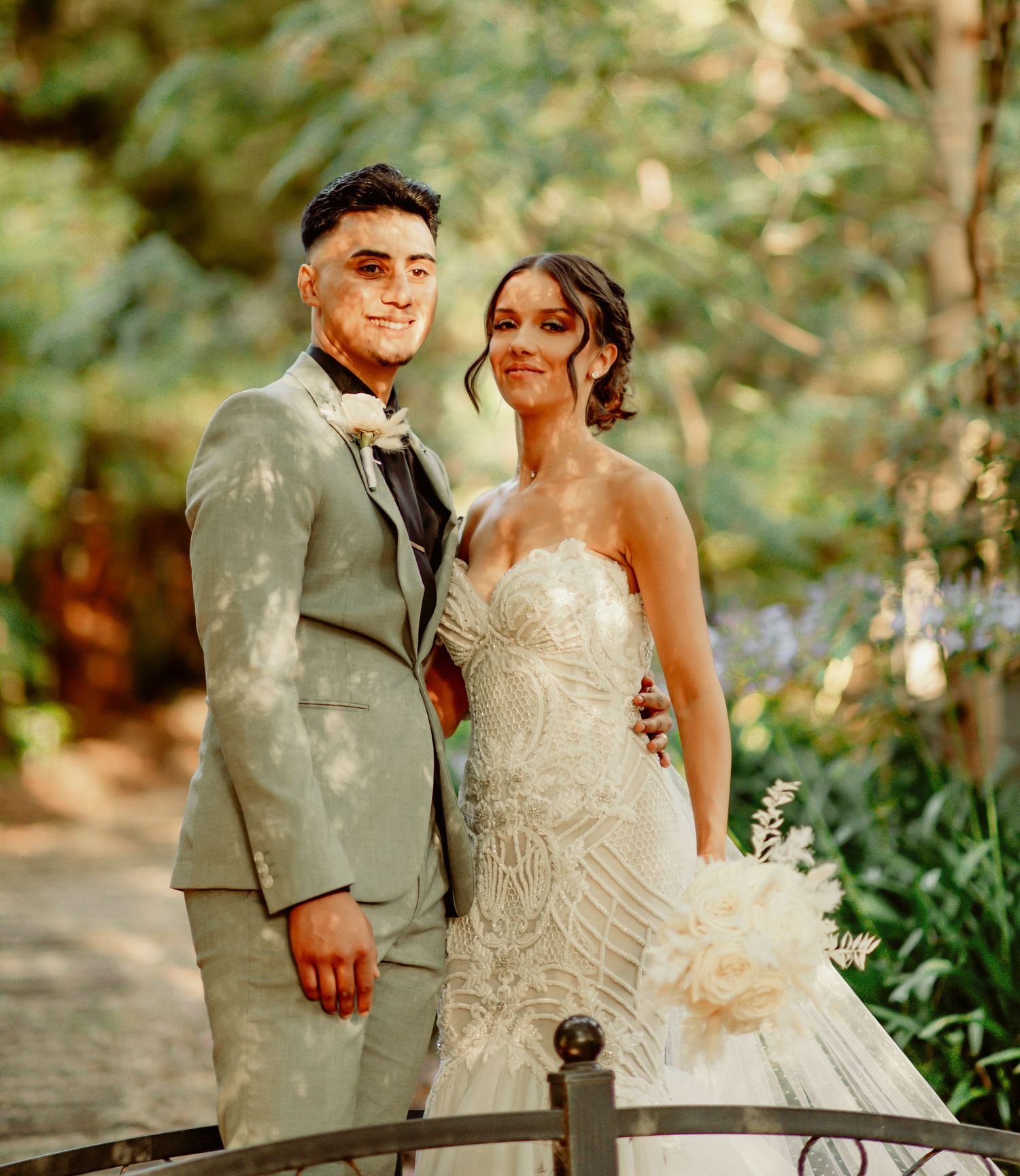 A newlywed couple poses together on their wedding day in a garden setting with natural sunlight filtering through trees.